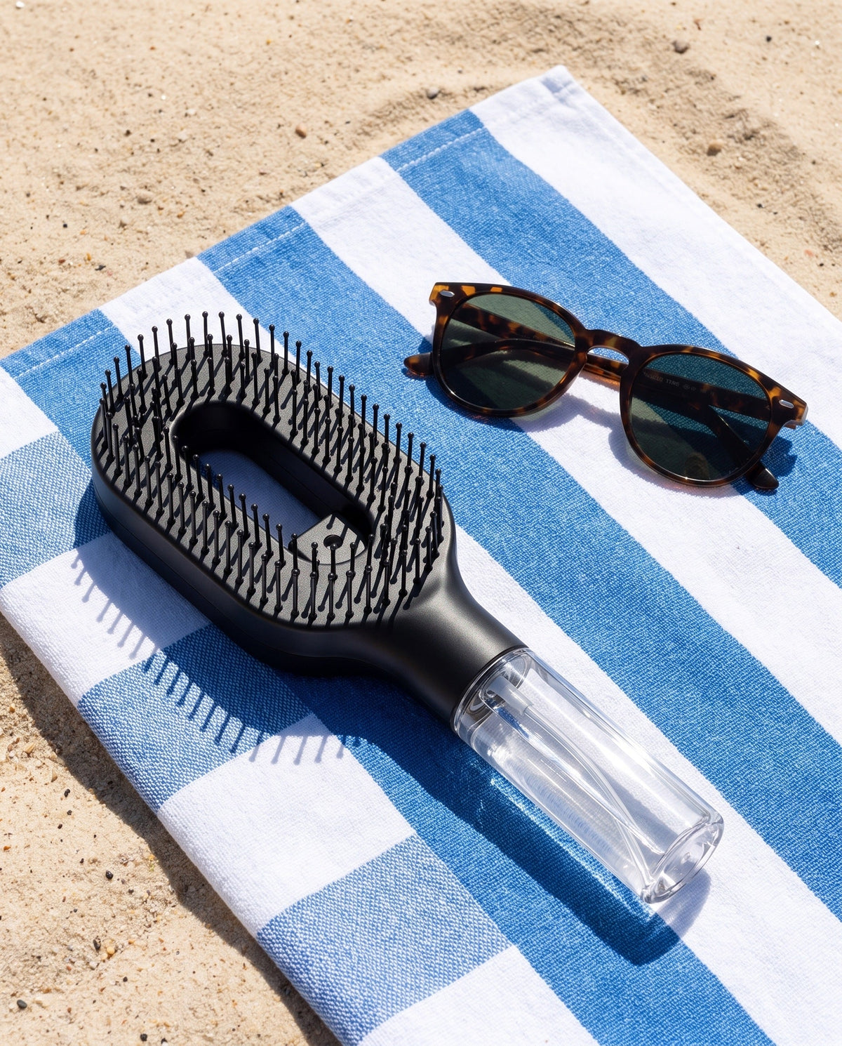 Brumelle Hydrating Brush in black on a striped beach towel in the sand beside sunglasses
