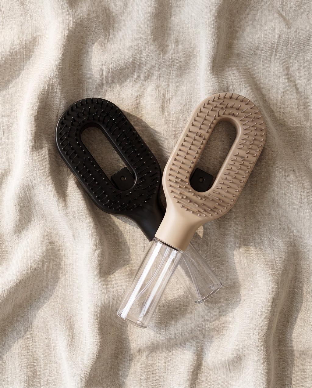The Brumelle Hydrating Brush in black and in beige crossed on linen, photographed from above with both clear refillable mist reservoirs visible